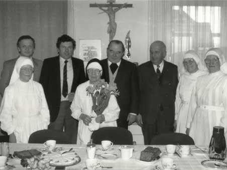 Schwester Brionda mit Blumen in der Hand und steht mit weiteren Personen hinter einem Tisch 